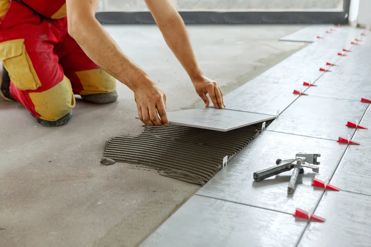 A person is kneeling and placing a Simple Placeholder Product 2 floor tile onto adhesive, surrounded by tools and spacers.
