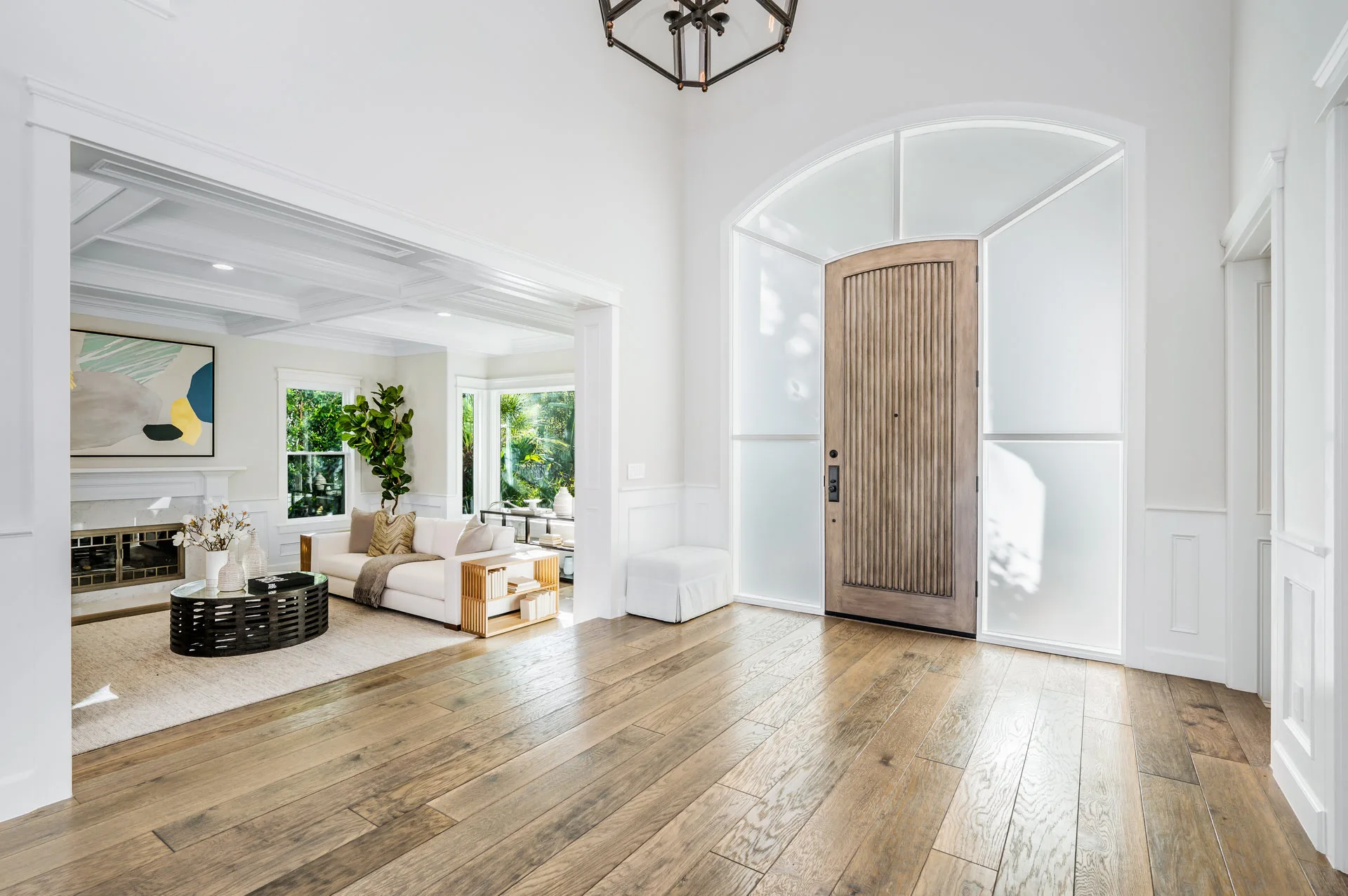 Bright entryway with wooden flooring, large arched door, and view of a living room with white couches, a modern coffee table, and artwork. Sunlight streams through windows, highlighting decor.
