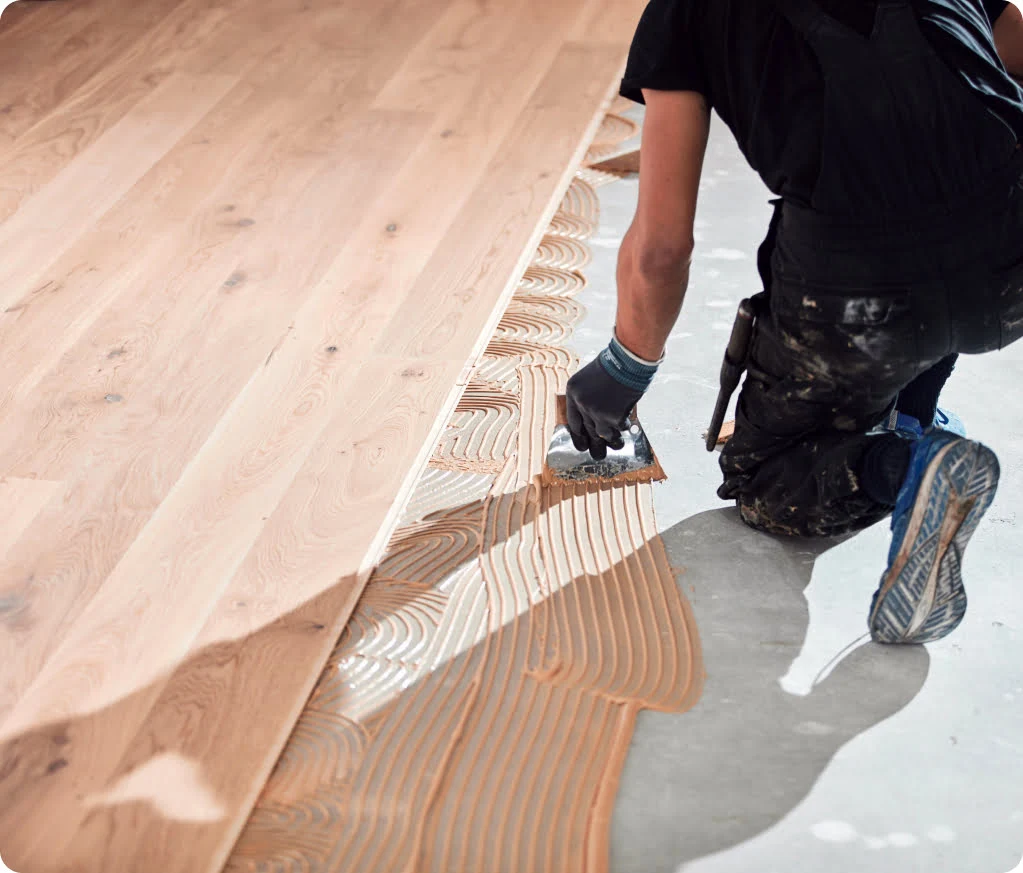 Person kneeling on the floor, applying adhesive in a wave pattern, preparing to install wooden flooring.