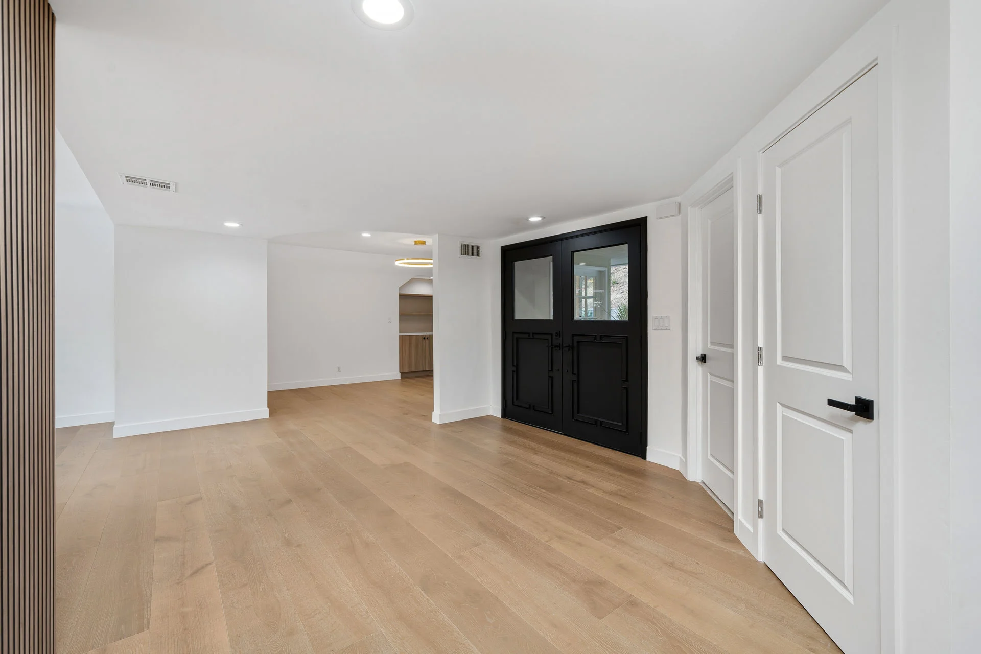 Spacious empty room with wooden floors, white walls, and a black double door. Recessed lighting illuminates the space.