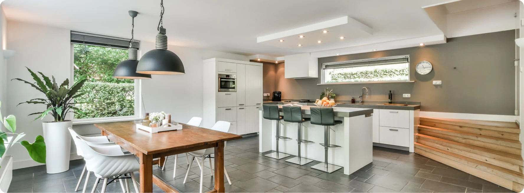 Modern kitchen interior with a wooden dining table, white chairs, large black pendant lights, and an island with barstools. Light gray tiled floor, white cabinets, and a window view of greenery.